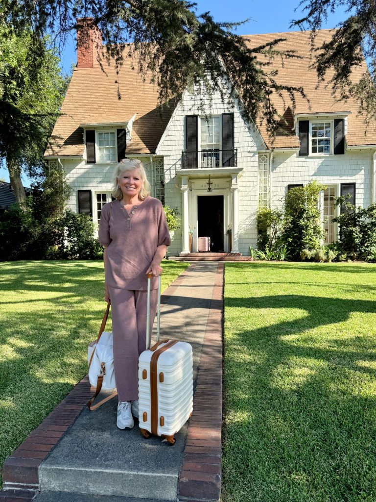 A woman wearing a matching set stands in front of a house, holding two suitcases. The house has a steeply pitched roof, dormer windows, and is surrounded by a well-maintained lawn.