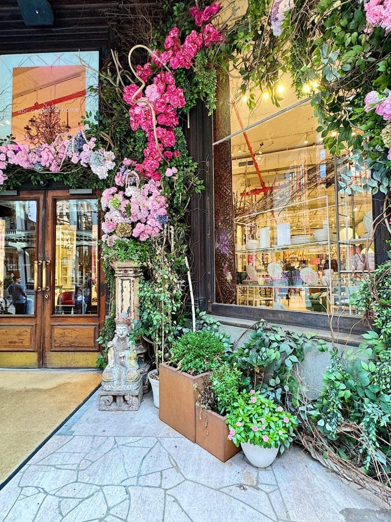 Entrance to a storefront adorned with abundant pink and purple floral decorations and lush green plants, with large windows displaying interior lighting and decor.