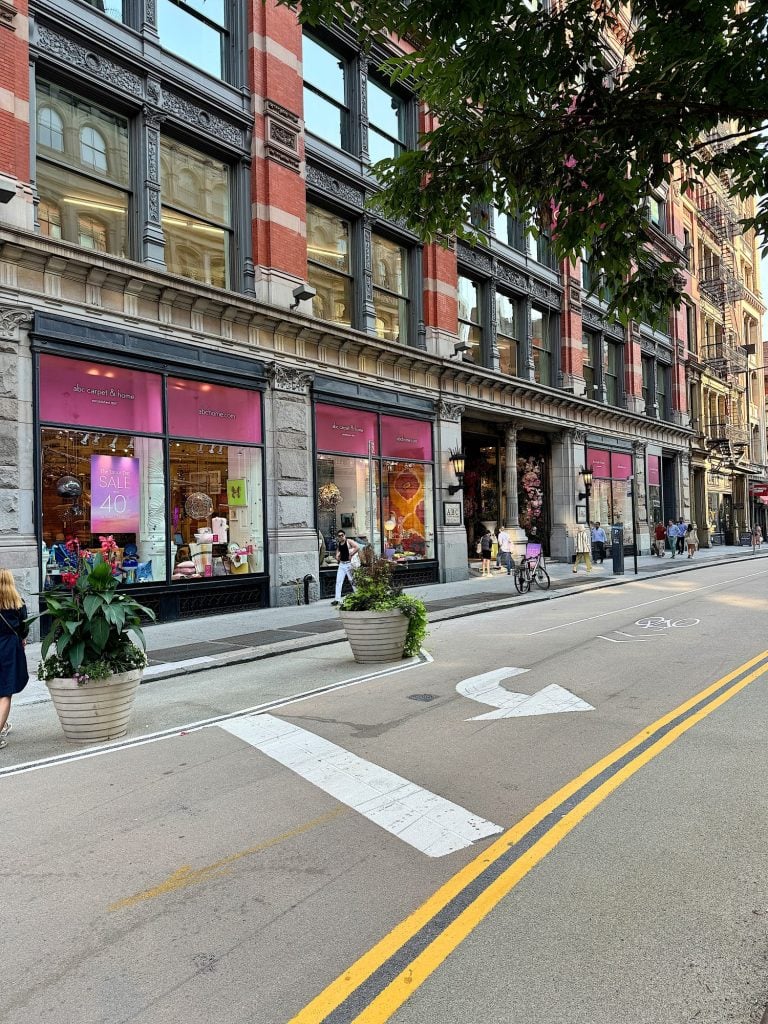 Street view of a building with pink window displays promoting a sale. Potted plants are placed along the sidewalk, and a few pedestrians and cyclists are on the street.