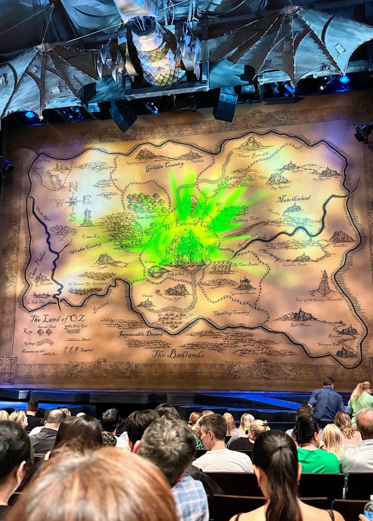 A theater audience faces a large, illuminated map of the fictional land of Oz on the stage backdrop, with green lighting prominently highlighting the center of the map.