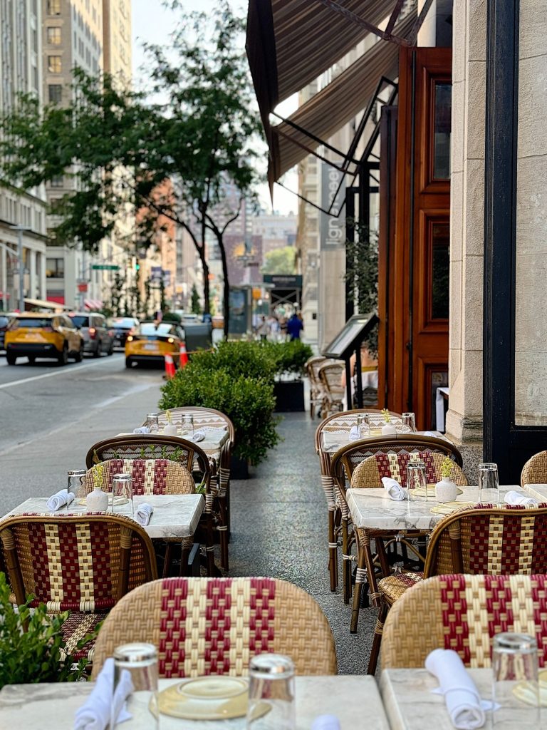 An outdoor cafe with empty tables and wicker chairs is set up along a city street with buildings, trees, and cars in the background.