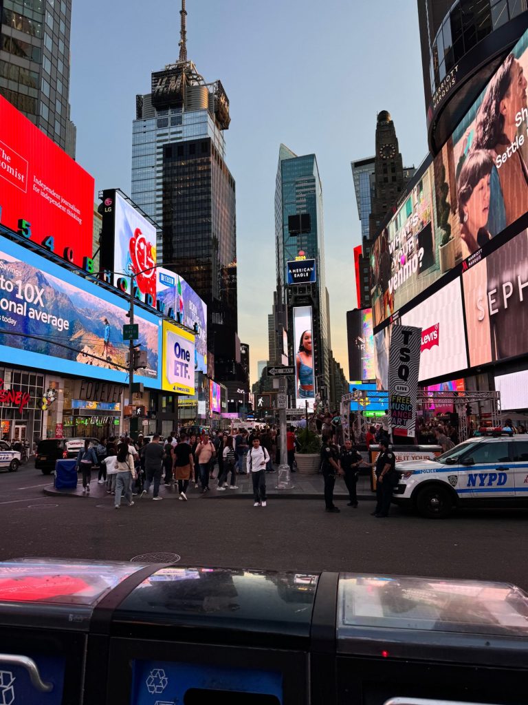 A bustling street in Times Square, New York City at dusk, filled with people and vibrant electronic billboards. Police cars are visible on the right side.