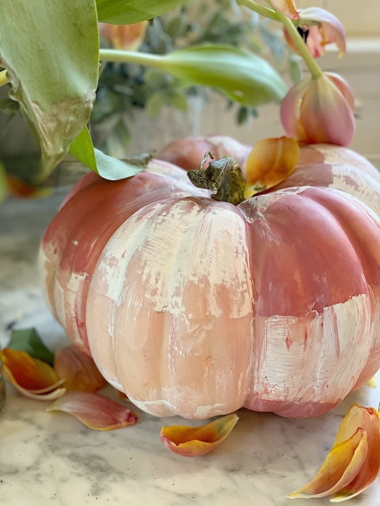 A painted pumpkin with a mix of pink and white colors sits on a marble surface, surrounded by scattered flower petals and green foliage.