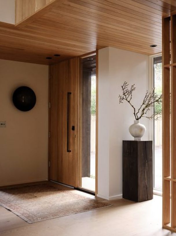 A modern entryway features a wooden front door, a tall wooden stand with a white vase holding branches, a round black wall decor, and a large rug on the floor.