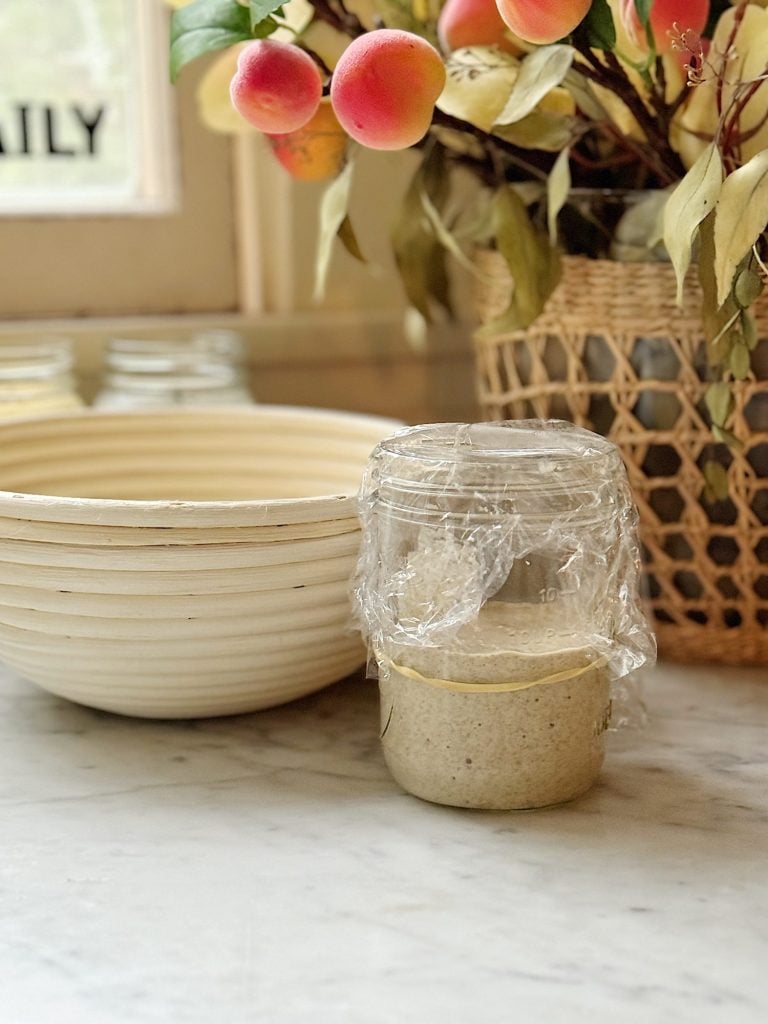 A glass jar covered with plastic wrap containing a dough-like mixture sits on a marble counter next to a beige bowl and a basket with artificial peach branches.