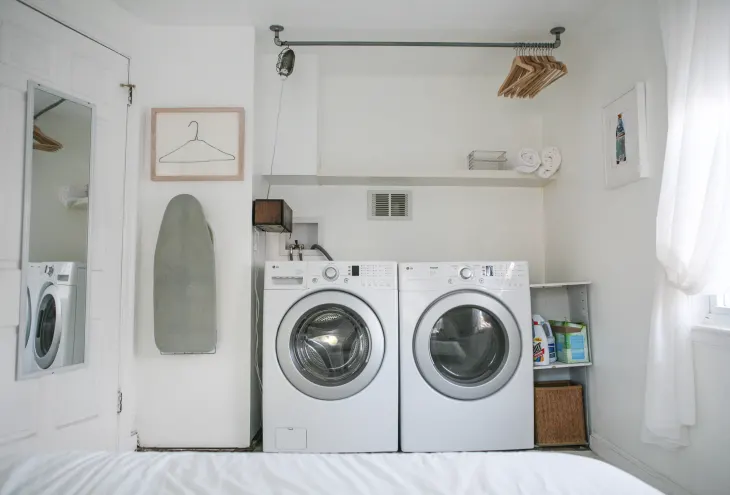 A white laundry room with a front-loading washer and dryer, ironing board on the wall, shelf with towels, and hangers.