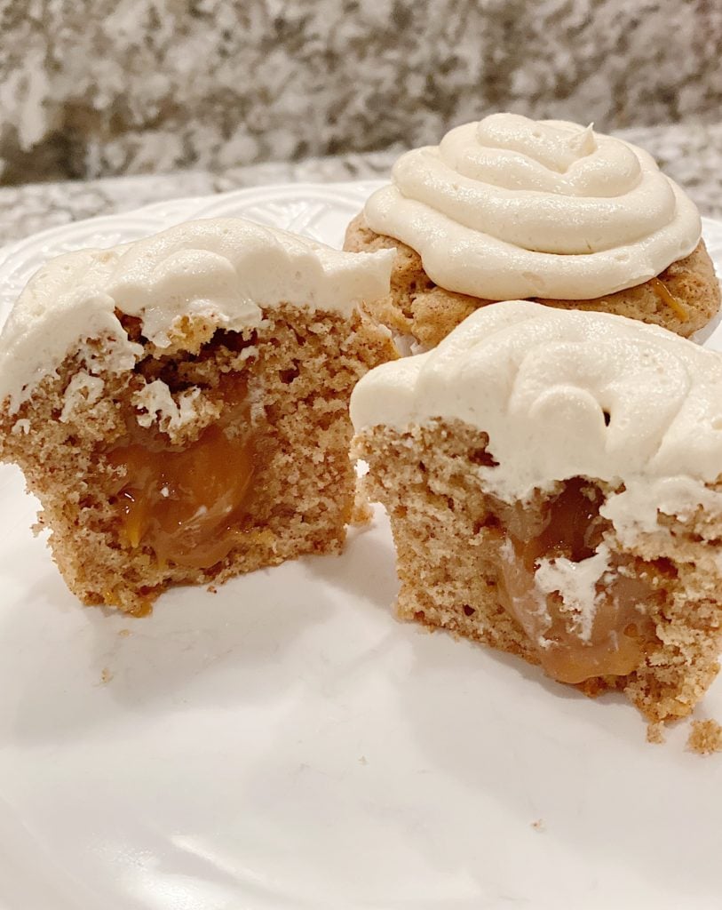 A sliced cupcake revealing caramel filling, topped with white icing, on a white plate with a textured background.