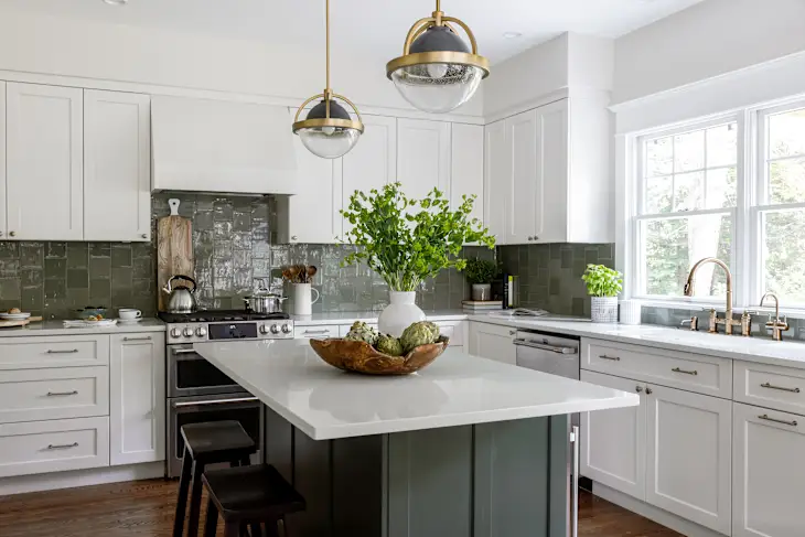 A modern kitchen with white cabinets, a central island with a white countertop, pendant lights, a stainless steel stove, and a plant centerpiece. The backsplash is a dark tile, and natural light enters through a window.