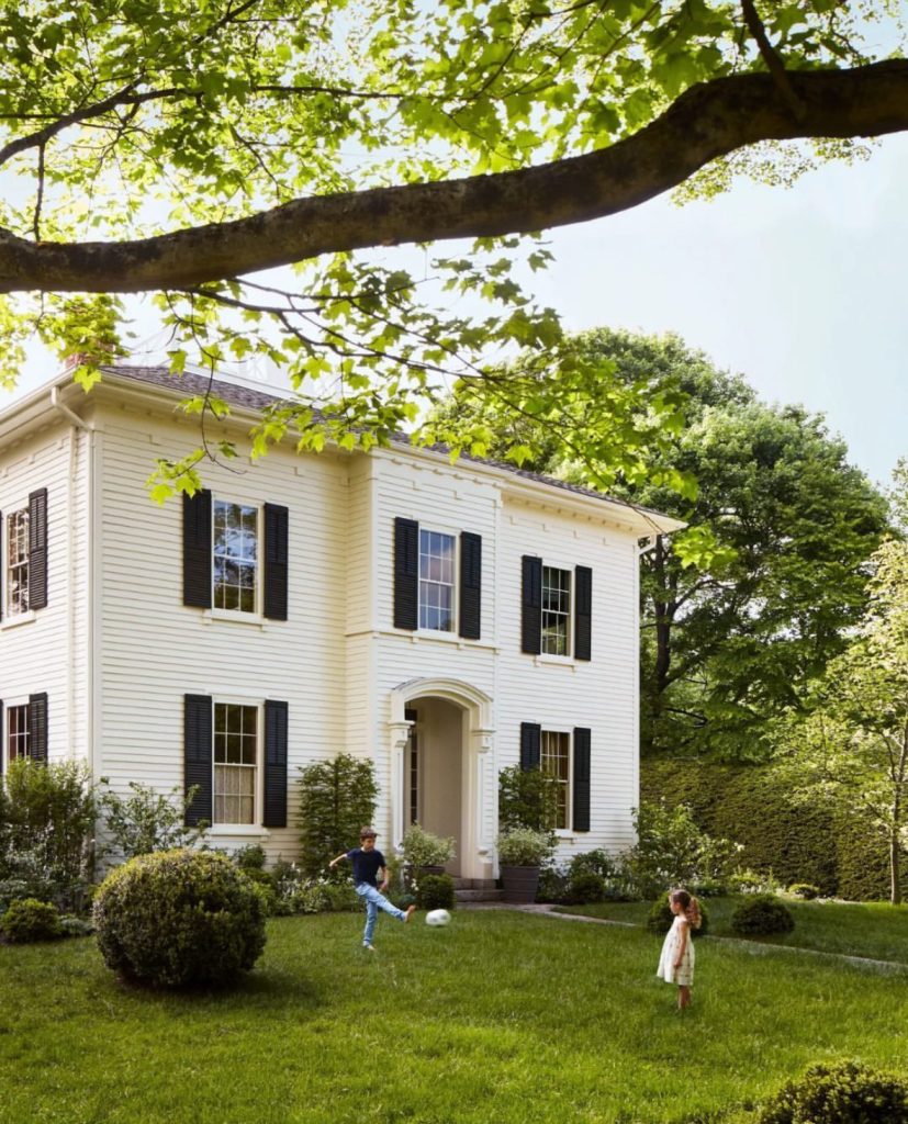 Two children play with a ball on the front lawn of a two-story white house with black shutters, surrounded by lush green trees and shrubs.