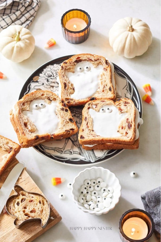 Three slices of ghost-themed toast with icing and candy eyes on a decorated plate. There are candles, small pumpkins, candy corn, and a bowl of additional candy eyes around the plate.