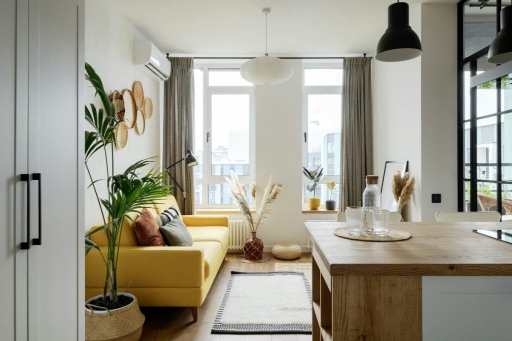 A modern living room with a yellow sofa, various houseplants, and neutral decor. Large windows allow natural light, and a wooden island is visible in the foreground.