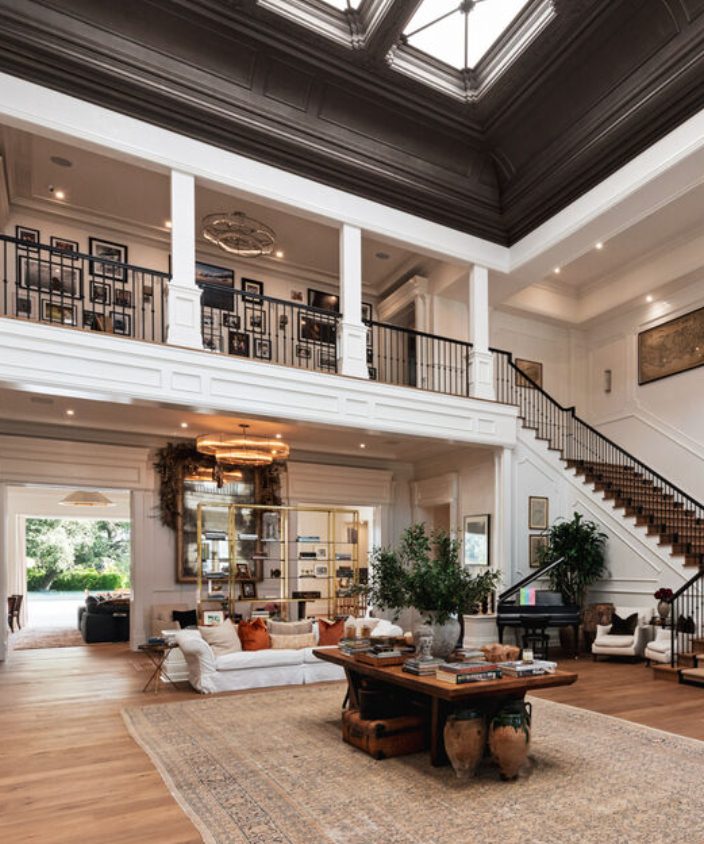 Spacious living room with high ceilings, a grand staircase, wooden floors, large rug, and various seating areas. Multiple framed pictures adorn the upper walls, while skylights let in natural light.