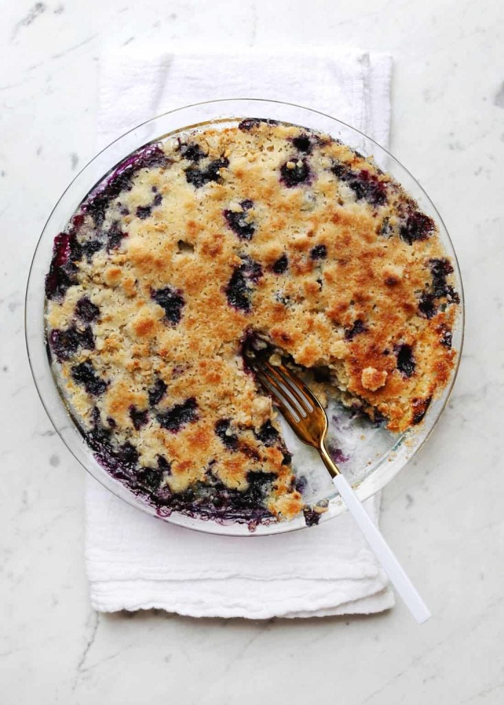 A round blueberry cobbler in a clear glass dish with one slice missing. A fork rests in the dish, and the cobbler is placed on a folded white cloth on a light-colored surface.