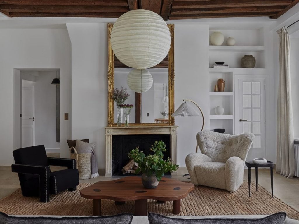 A modern living room with two armchairs, a coffee table, a large mirror above a fireplace, shelves with decorative items, and a paper lantern pendant light.