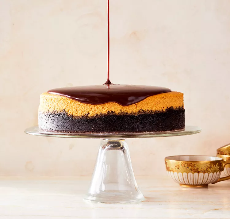 A layered cake with caramel and chocolate being drizzled with chocolate sauce on a glass cake stand. Several ornate bowls are placed nearby.