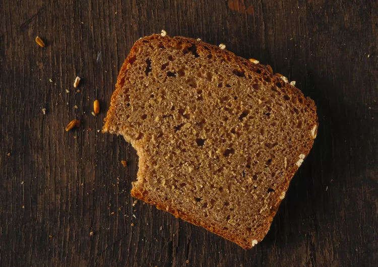 A brown slice of bread with a bite taken out, placed on a dark wooden surface with some crumbs around it.