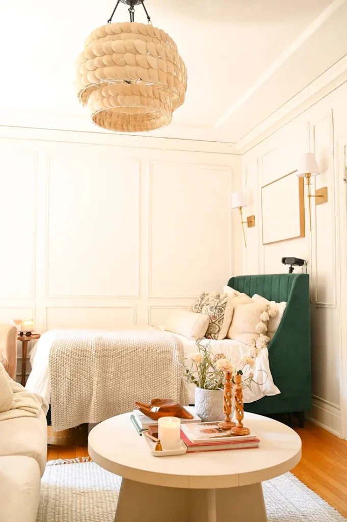 A cozy bedroom with a green upholstered bed, round coffee table with decor items, white walls, and a unique layered chandelier.