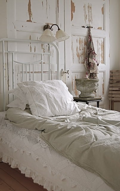 A white metal bed with a lace bedspread and beige bedding against a distressed white-wood wall, with a wooden side table, a lamp, and a potted plant.