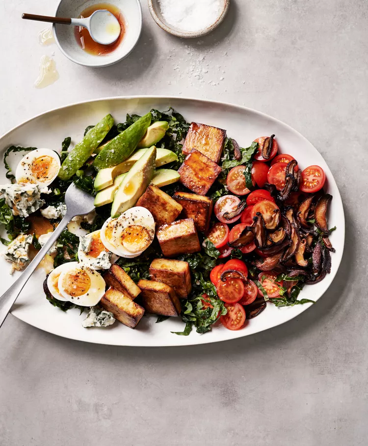 A plate of food featuring roasted tofu, hard-boiled eggs, avocado slices, mushrooms, cherry tomatoes, and mixed greens, accompanied by a small bowl of sauce and a bowl of salt on the side.