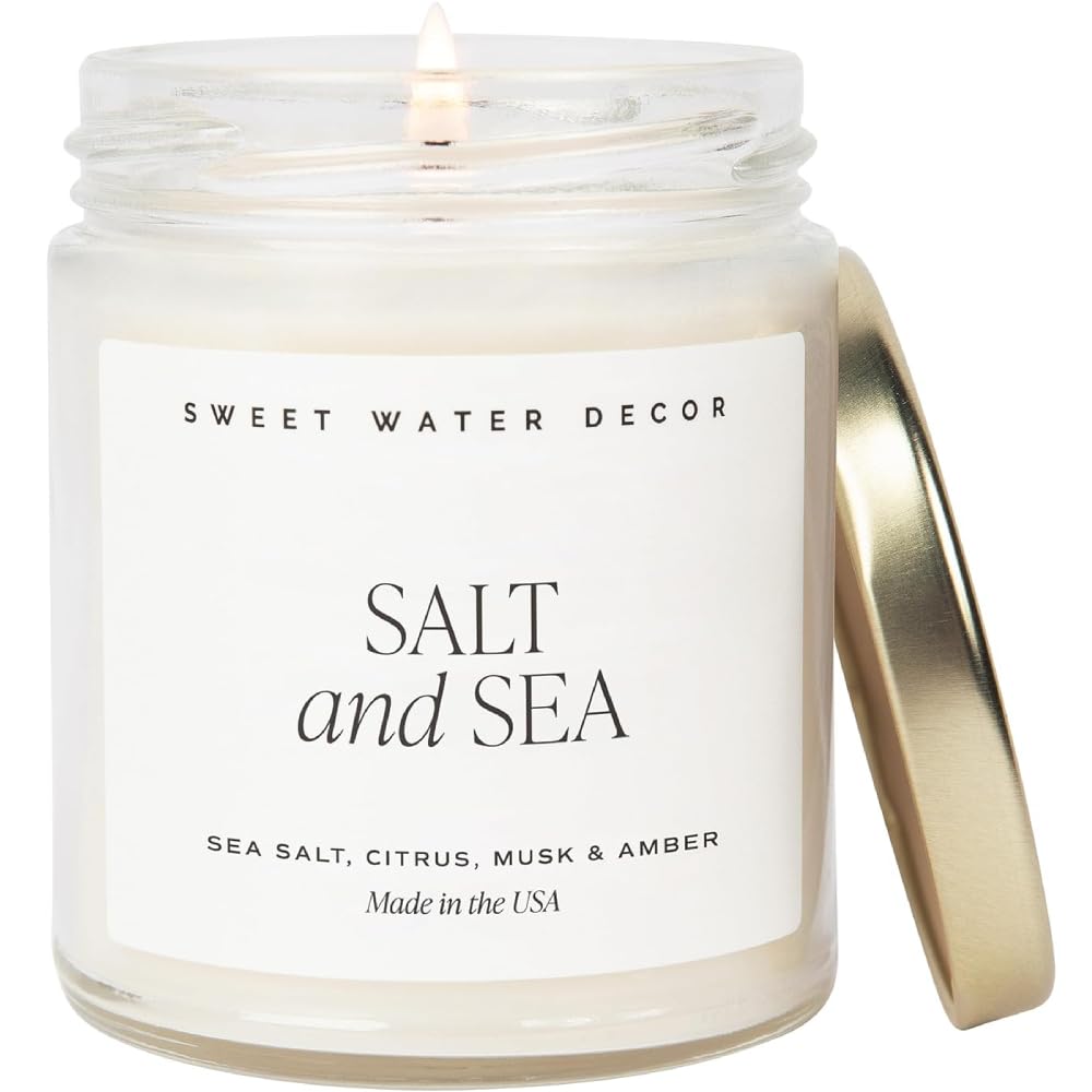 A candle labeled "Salt and Sea" by Sweet Water Decor, featuring scents of sea salt, citrus, musk, and amber. The lid is off and a flame is lit. Made in the USA.