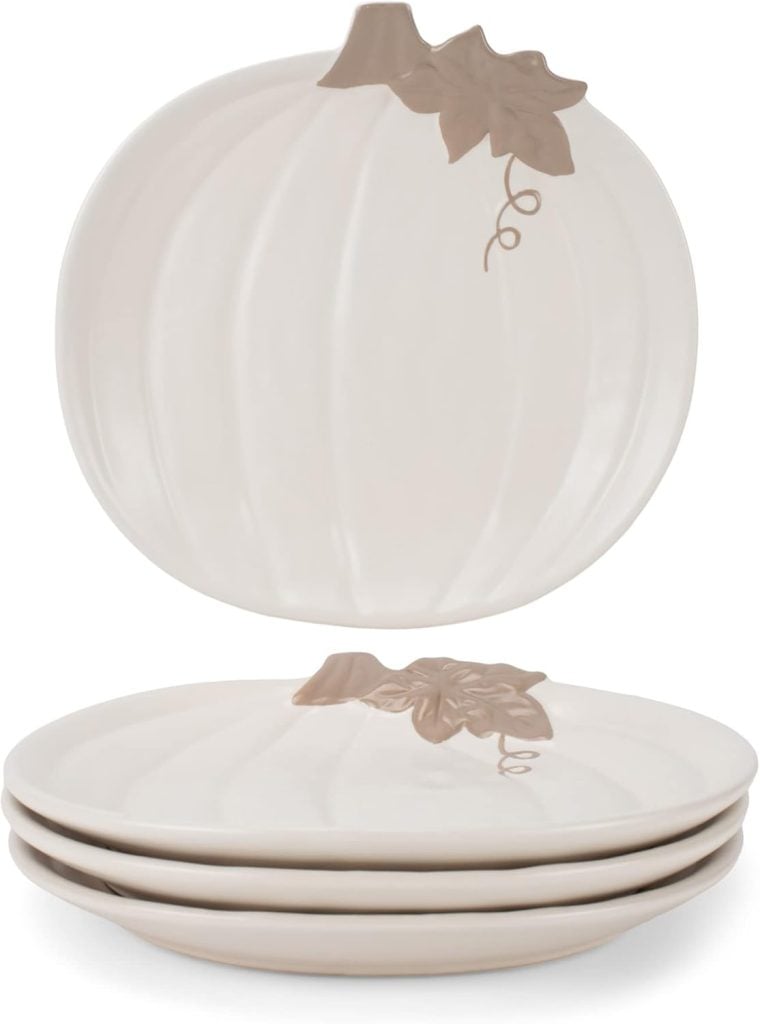 Four cream-colored, pumpkin-shaped Thanksgiving plates with brown leaf detailing are stacked, with one plate standing upright behind them.