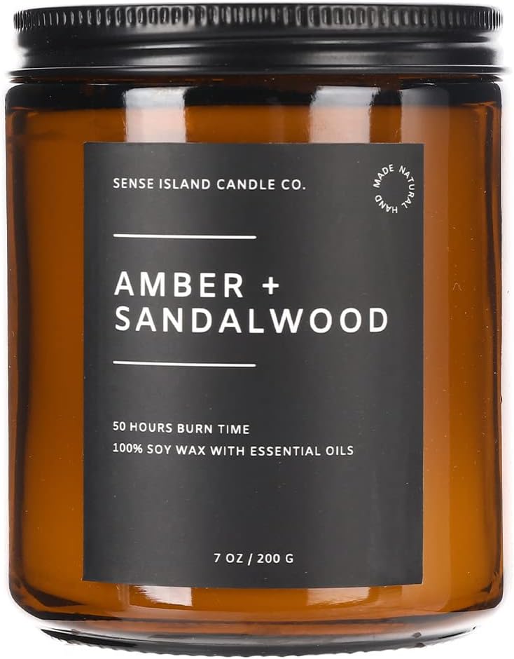 Brown glass candle jar with a black label reading "Amber + Sandalwood" from Sense Island Candle Co. It states "50 hours burn time" and "100% soy wax with essential oils.