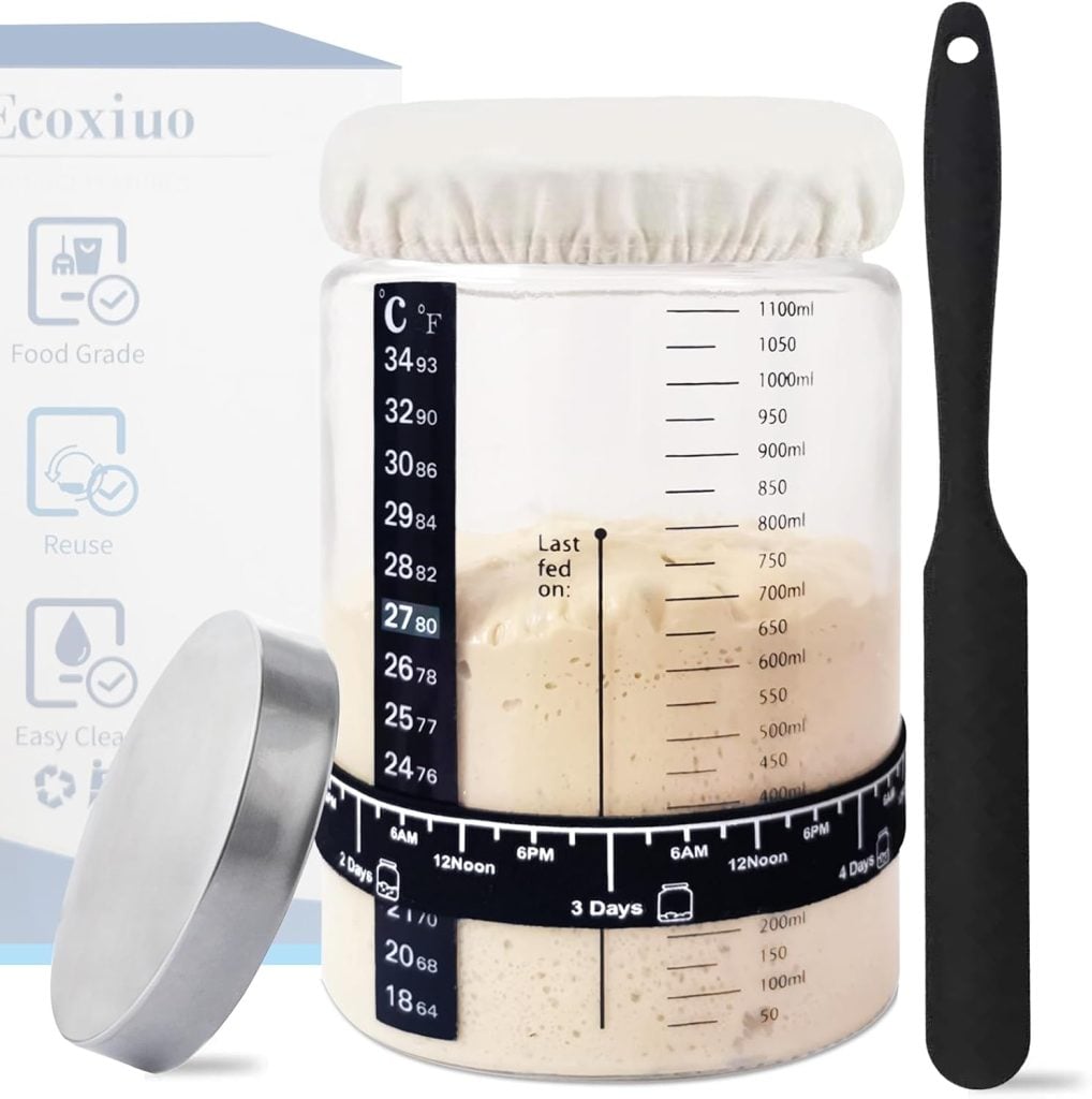 A glass jar with measurement markings and a cloth cover contains dough, illustrating how to make a sourdough starter; a black spatula and a metal lid sit nearby, with product information featured in the background.