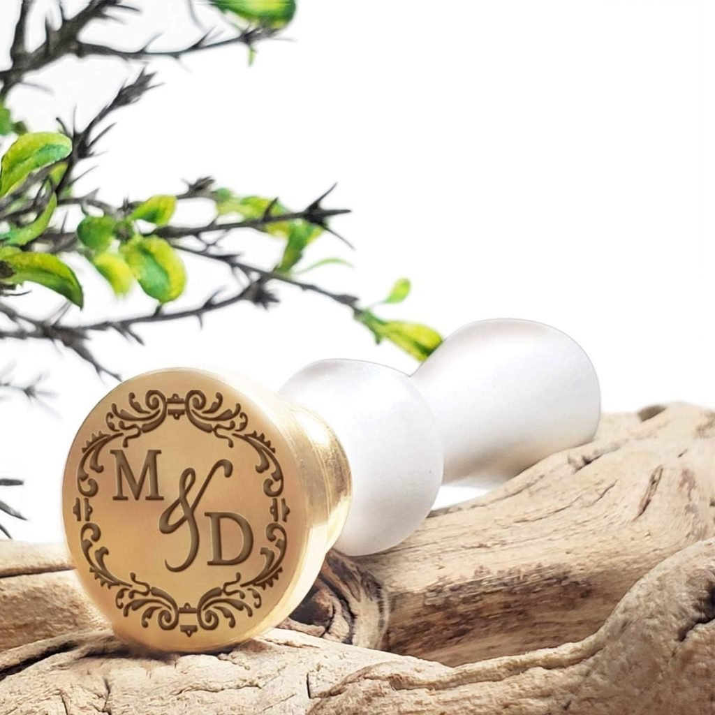 A gold wax seal stamp with intricate initials "M & D" rests on a piece of wood beside green leaves.