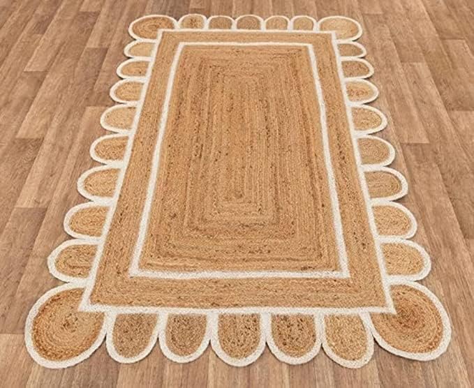 A rectangular jute rug with a scalloped edge lies on a wooden floor. The rug features a two-tone design with a darker center and lighter-colored edges.