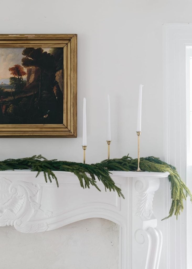 White fireplace mantel with green garland, three tall white candles in gold holders, and a framed landscape painting above.