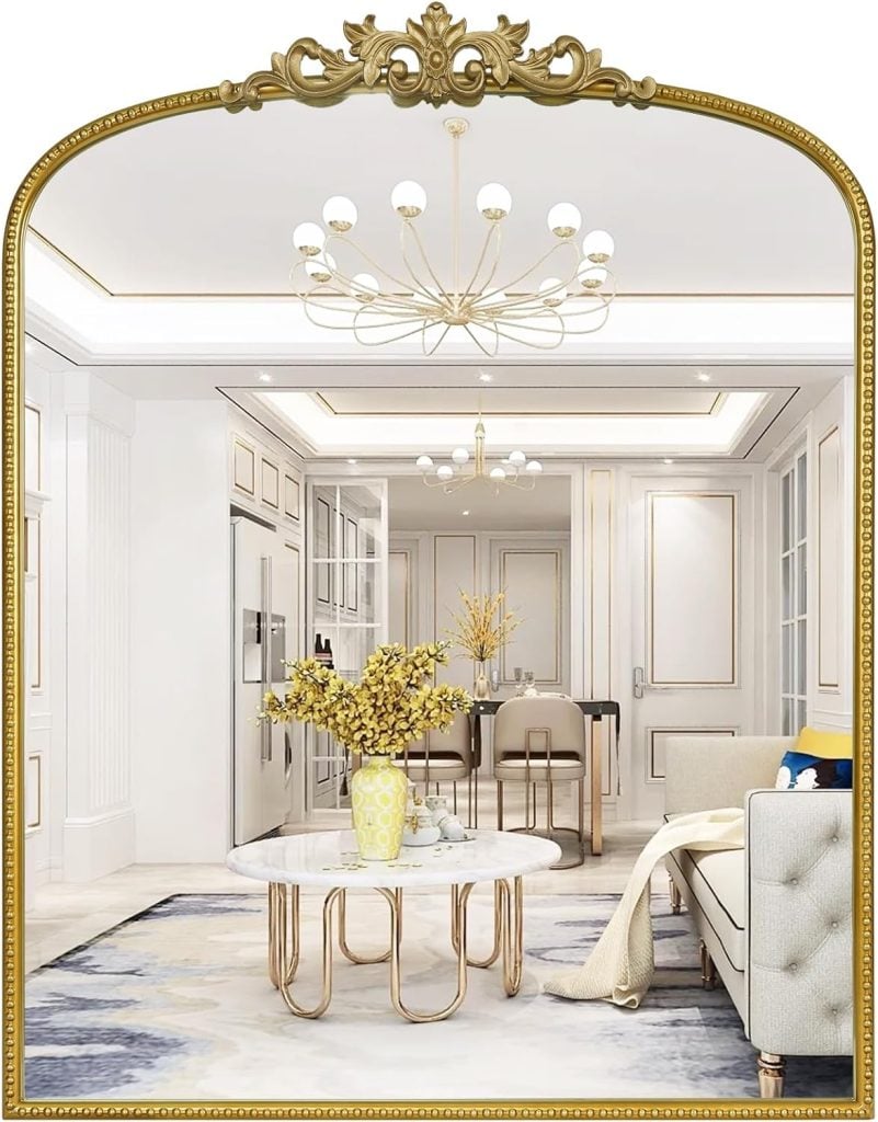 Elegant living and dining area with a gold-framed mirror, chandelier, marble coffee table, beige sofa, and vase of yellow flowers.