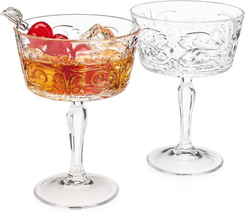 Two ornate glass goblets sit next to an array of Thanksgiving plates; one contains an amber liquid with ice and cherries, while the other remains empty.