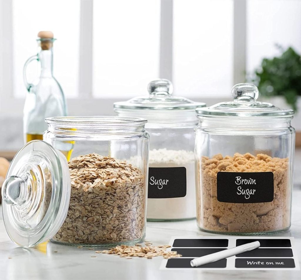 Three glass jars, labeled "Sugar," "Brown Sugar," and an unlabeled one with oats, sit on a kitchen counter. A partially visible bottle and some green foliage are in the background, evoking the cozy atmosphere of preparing Baked Brie with Jam.