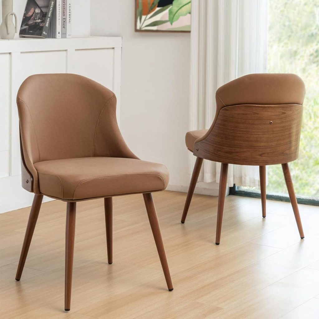 Two brown upholstered chairs with wooden legs are positioned on a light wooden floor near a window with sheer white curtains. A white cabinet and plant picture are in the background.