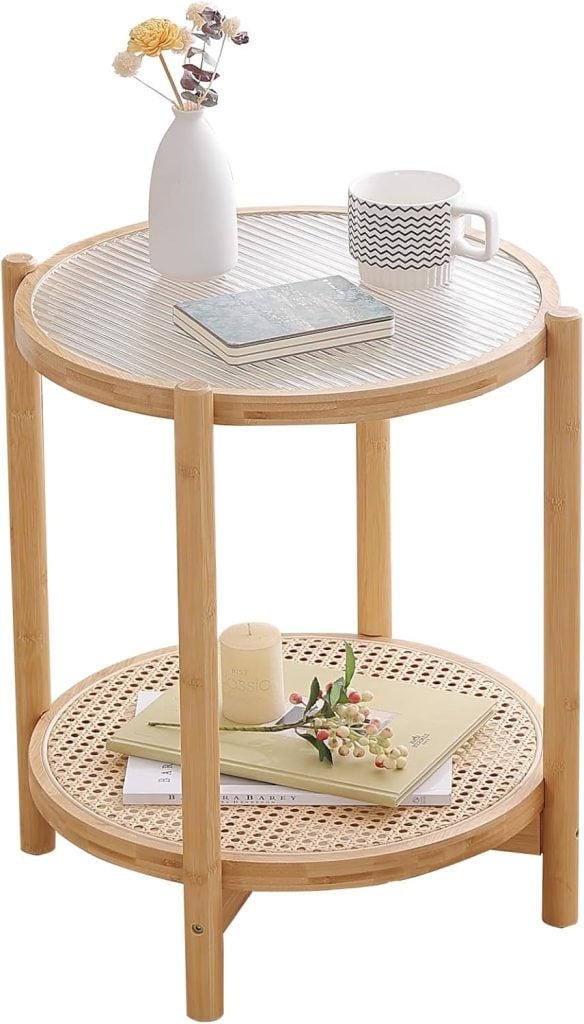 A small, round, two-tiered wooden table holds a vase with flowers, a book, and a coffee cup on the top tier, with a candle and additional book on the bottom tier.