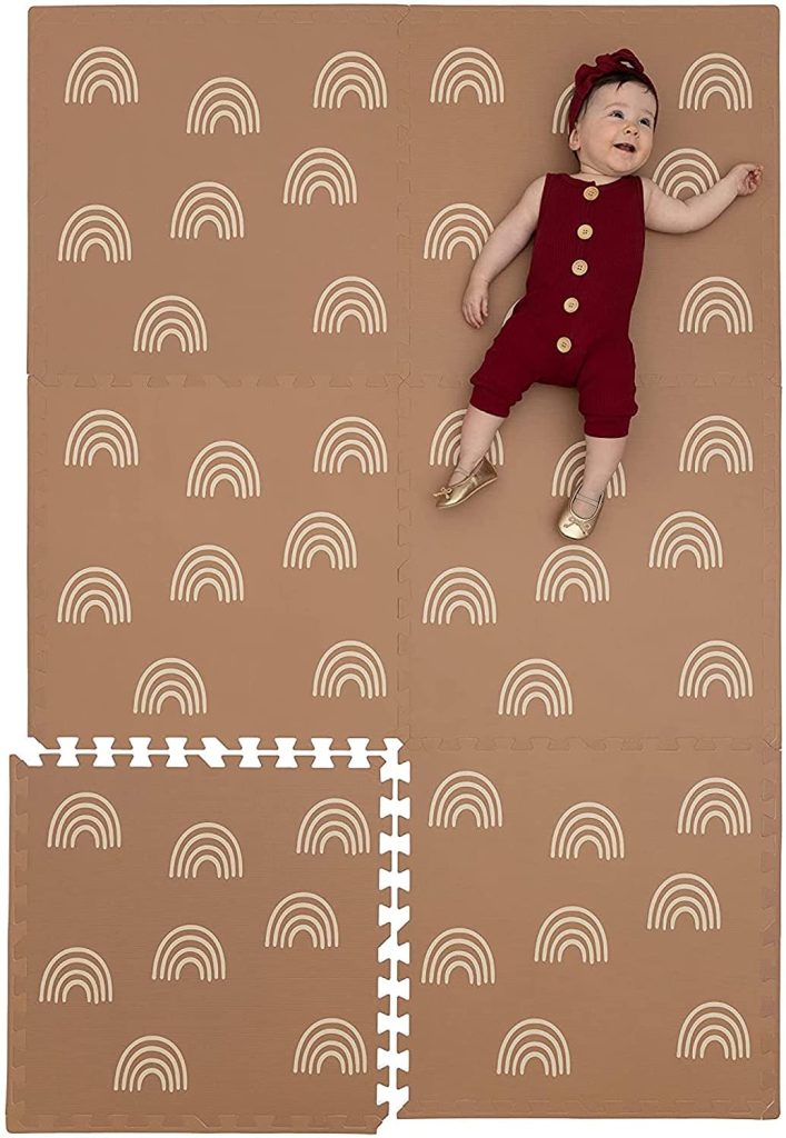 A baby in a red outfit lies on brown foam play mats adorned with white rainbow patterns, creating a picture-perfect moment that could easily grace an Amazon Prime family ad.