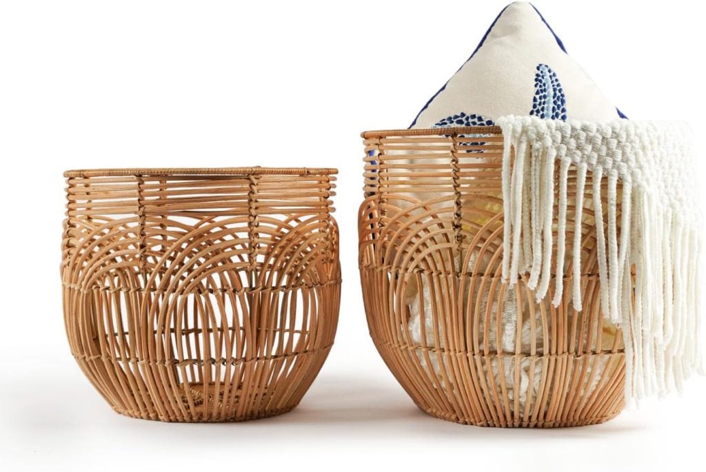 Two woven rattan baskets, one empty and one containing a white pillow with blue design and a white fringed blanket, are placed on a white background.