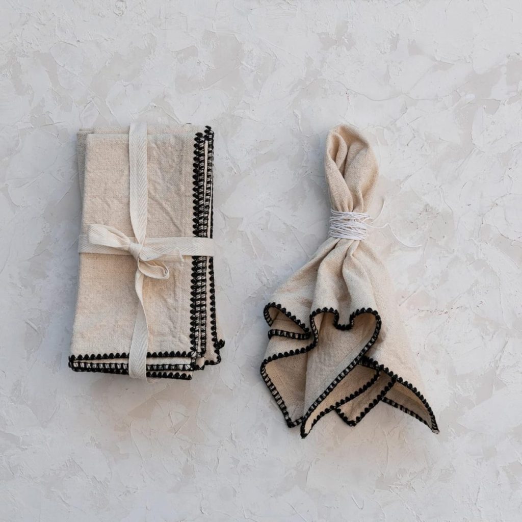 Two beige linen napkins with black trim—one folded and tied with a ribbon, the other gathered at the center—rest on a textured white surface, perfectly suited for cozy fall soup recipes.