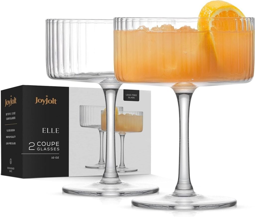 Two JoyJolt coupe glasses, one filled with an orange drink and a citrus slice, are displayed alongside their packaging box. Available for quick delivery with Amazon Prime.