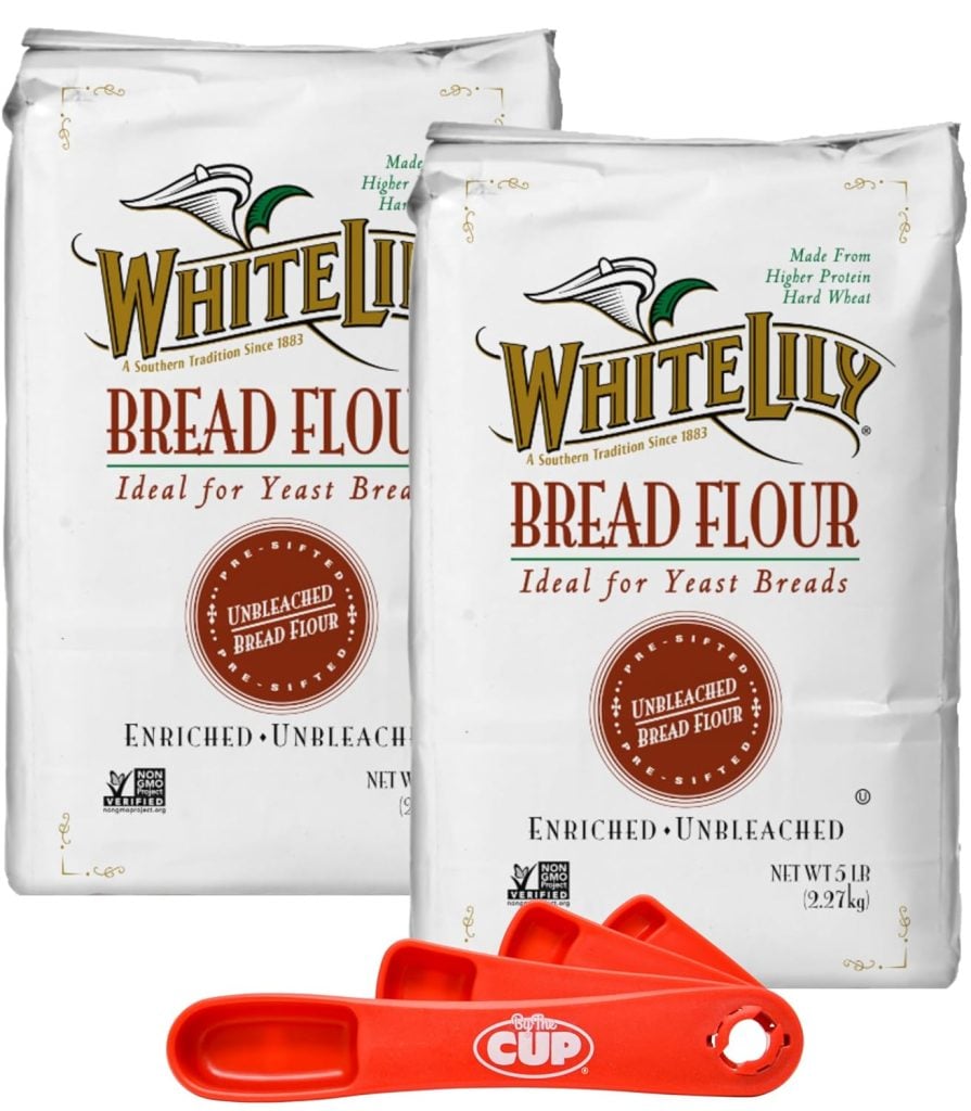 Two bags of White Lily bread flour labeled "enriched unbleached," with red measuring cups in front.