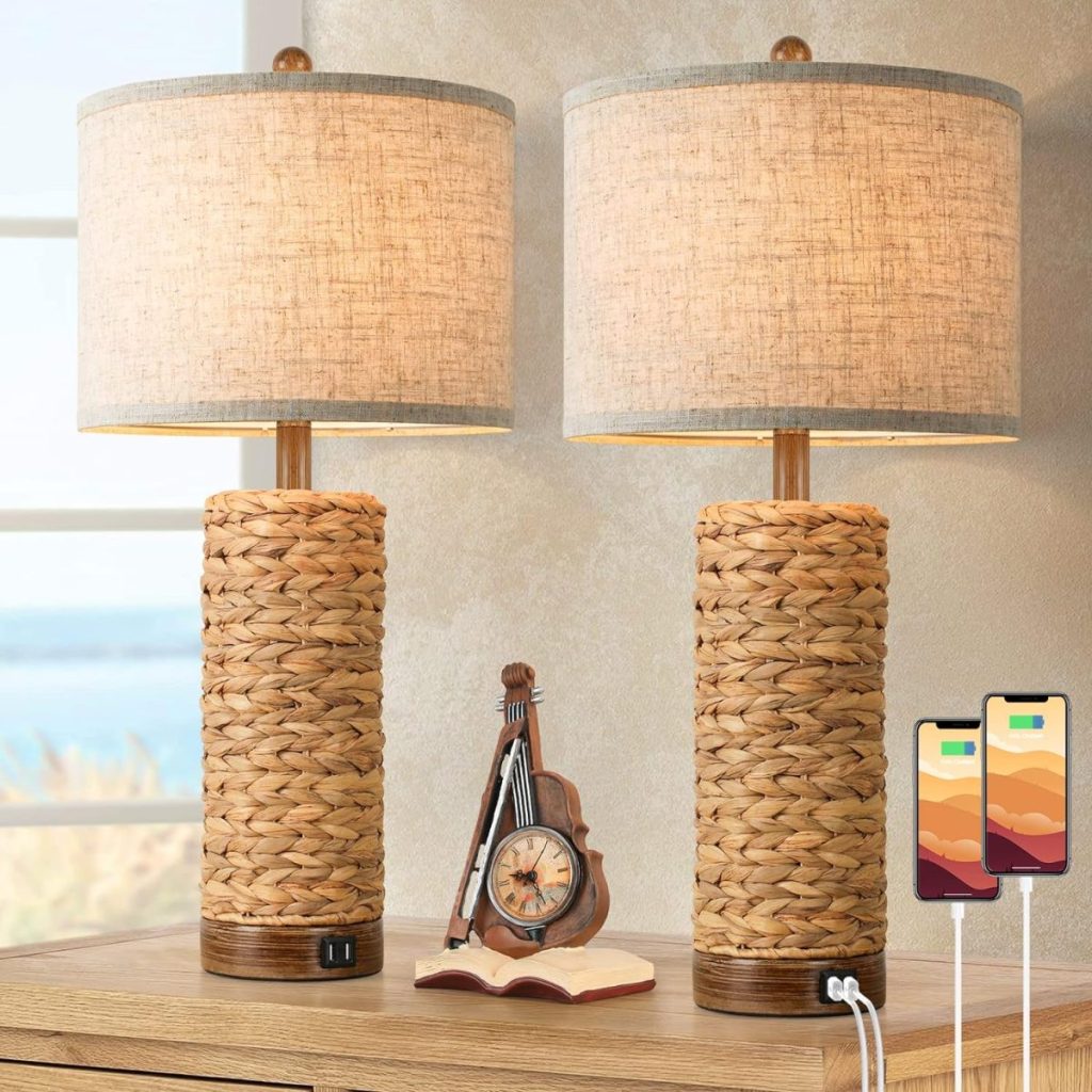 Two woven table lamps with beige shades on a wooden surface, flanking a small clock and a miniature violin. Two smartphones are plugged into USB ports at the base of one lamp.