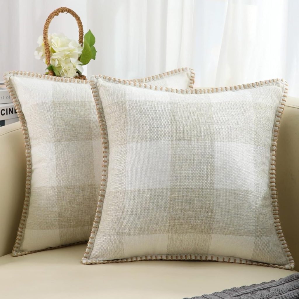 Two beige checkered pillows with braided edges are placed on a light-colored sofa. A book and a vase with white flowers are in the background.