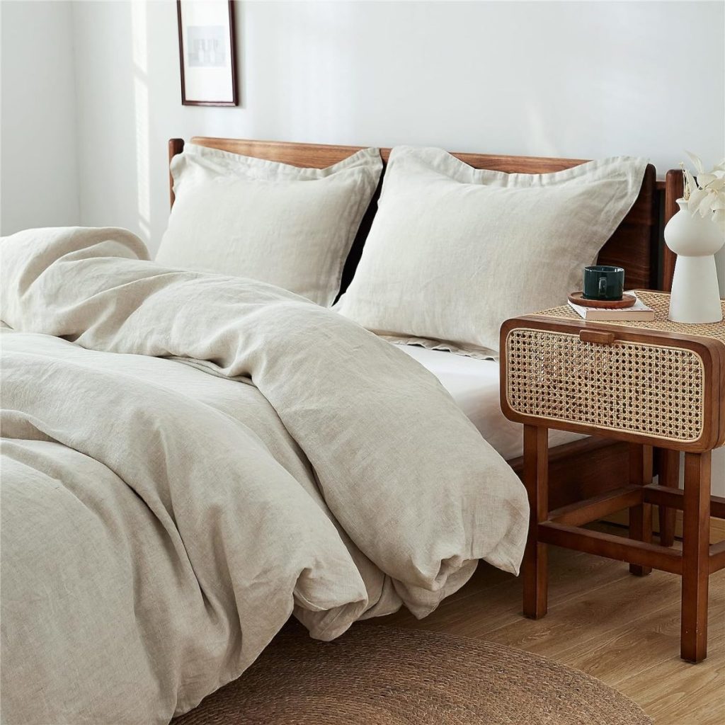 A neatly made bed with beige linen bedding, wooden frame, and two pillows. A wooden nightstand with a wicker front holds a small vase and a cup. A framed picture hangs on the wall above the bed.