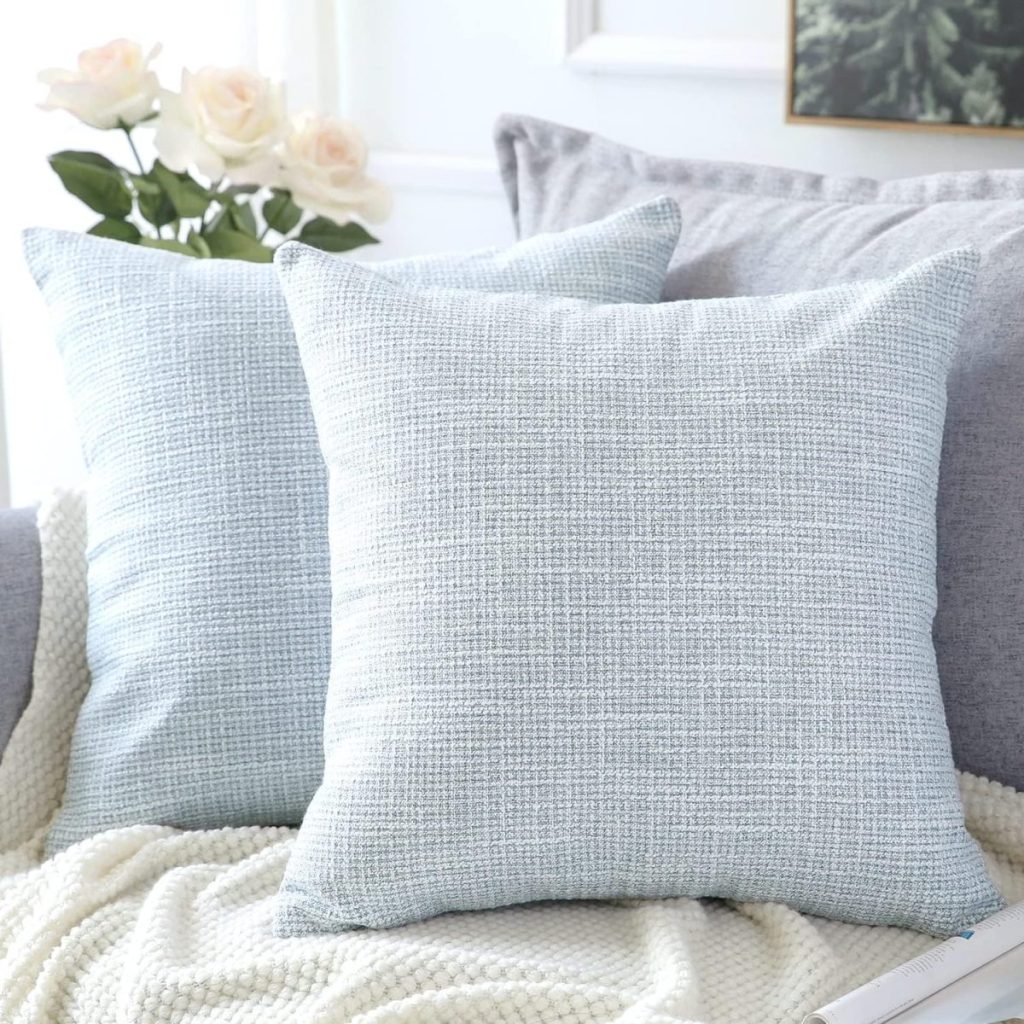 Two light blue textured pillows are placed on a light gray couch with a white knitted blanket. In the background, there are white flowers in a vase and a partially visible framed picture.