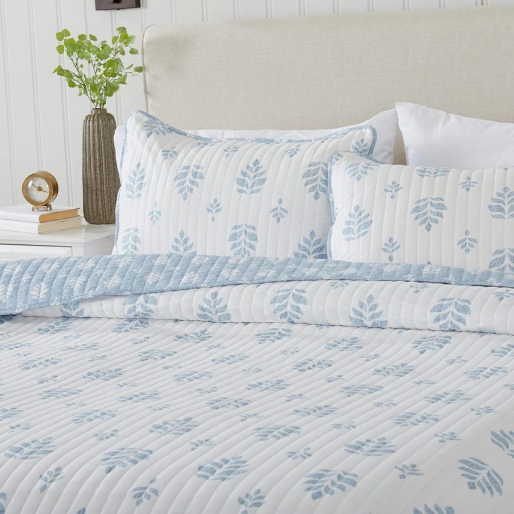 Bed with blue and white floral patterned bedding, including pillows and quilt, next to a white nightstand with a plant, clock, and stack of books.