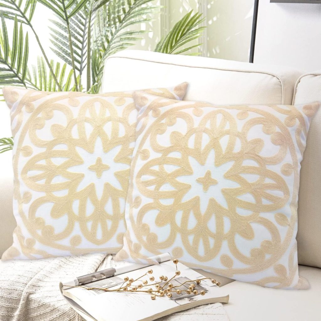 Two decorative pillows with beige and white floral patterns on a light-colored sofa, surrounded by green plants.