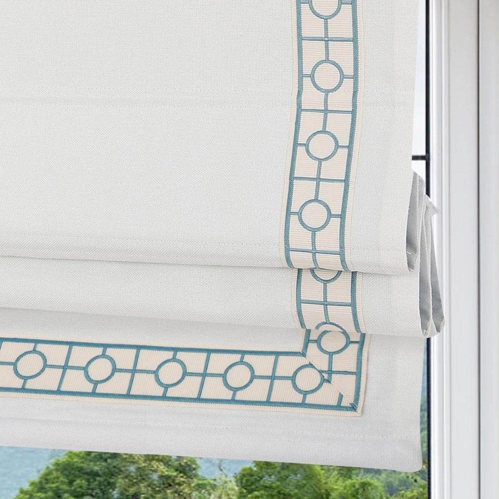 Close-up of a white roman shade with blue geometric trim, partially raised, with a view of green tree foliage through the window in the background.