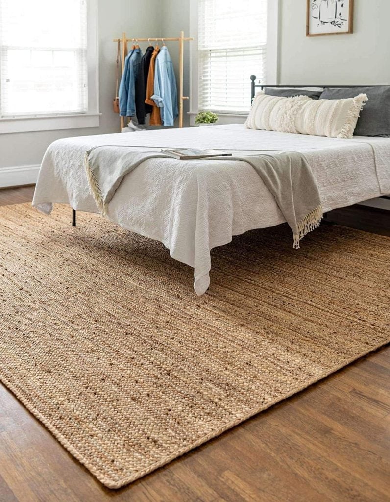 A bedroom with a bed covered in white and beige bedding, a jute rug on a wooden floor, clothing rack in the corner, and two large windows with blinds.