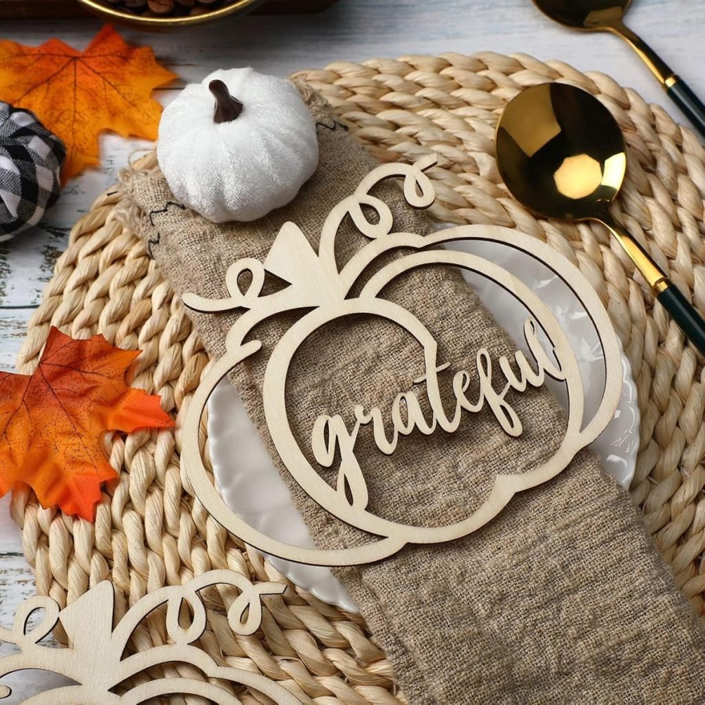 A wooden "grateful" decoration in the shape of a pumpkin sits on a woven placemat, perfect for Friendsgiving ideas, accompanied by a gold spoon and fork, a small white pumpkin, and a fall leaf.