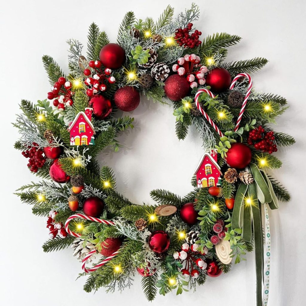 Festive Christmas wreath with red baubles, berries, greenery, candy canes, and small house ornaments, adorned with lights and a ribbon.